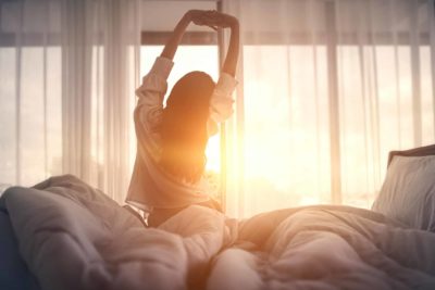 Woman stretches as she gets out of bed facing the morning sun, showing how sleep issues may be a sign of anxiety.