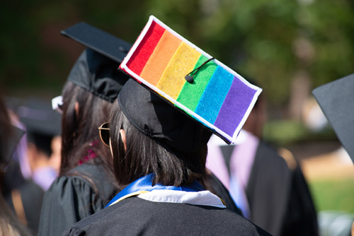 Woman wears a black graduation gown, with a rainbow-striped graduation cap.
