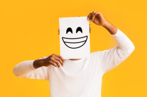 Black man holding paper with smiley face printed on standing in front of orange background signaling high functioning depression