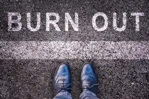 Arial picture of word “burn out” written in in white on black asphalt with men dress shoes of healthcare professional.