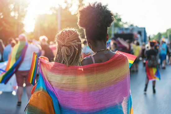Young, female Black couple hugging with a rainbow scarf wrapped around them both watching the sunset at a Pride event
