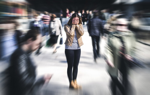 Woman with her face in her hands having a panic attack, while blurred images of people rush around her.