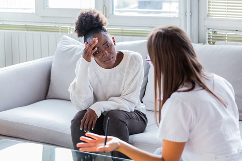 Frustrated Black patient sitting on couch next to white mental healthcare provider in therapy session