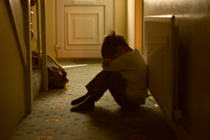 Young child sitting on the floor in a dark hallway with his arms wrapped around knees, indicating depression