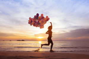 Woman running on beach at sunset, holding a bundle of red, orange and yellow balloons, signifying good mental health.
