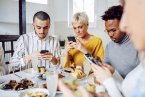 Group of multicultural friends sitting at dining table for lunch and using smartphones showing how technology can impact mental health.