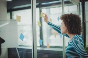 Black woman writing a sticky note on a glass wall showing 5 facts to know about mental health