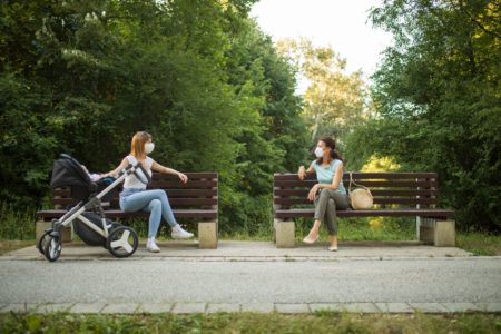 Two women talk on park benches 6’ apart while one mom keeps a hand on her baby’s stroller