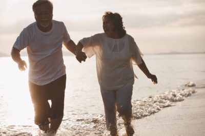 Senior Asian couple frolics on beach at sunset, illustrating that anxiety while aging can and should be treated.