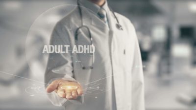 Cropped photo of male doctor holding out hand with the words “ADULT ADHD” circa imposed on the frame.