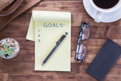 Picture from above of a small yellow notepad that says “Goals” and three action items, with a pen laying on top, illustrating importance of setting therapy goals.