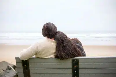 Biracial couple leans on each other on bench at beach, navigating grief during the holidays.