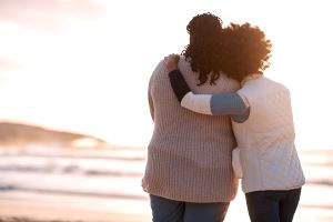 Two black women hug on a beach looking at a sunset, illustrating importance of connection during depression