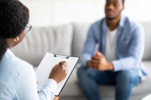 Young black guy with depression or PTSD having session with psychotherapist at clinic, selective focus. Sad African American man having emotional problem, receiving professional psychological help