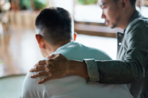 Rear view of son and elderly father sitting together at home. Son caring for his father, putting hand on his shoulder, comforting and consoling him over his mental health