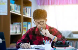 young asian boy wearing eyeglasses and rainbow wristband, holding pen, sitting in library, reading books and concentrating indicating the importance of caring for LGBTQI+ mental health