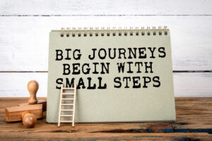 Big Journeys Begin With Small Steps. Green notepad on wooden texture table and white background representing creating small habits to benefit your mental health