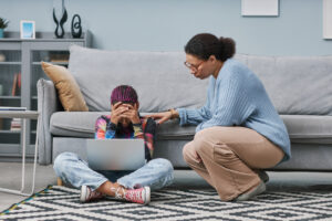 Young woman of color with hand covering face and laptop in lap being consoled by mother over her anxiety because of cyberbullying