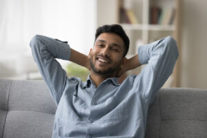 Happy attractive young Indian guy enjoying leisure, comfort at home, relaxing on comfortable sofa, stretching body, leaning on back, looking at camera with toothy smile. 