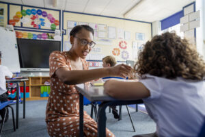 African American woman teacher looking concerned crouching next to a young student whose head is down on the desk indicating anxiety and behavior changes from going back to school