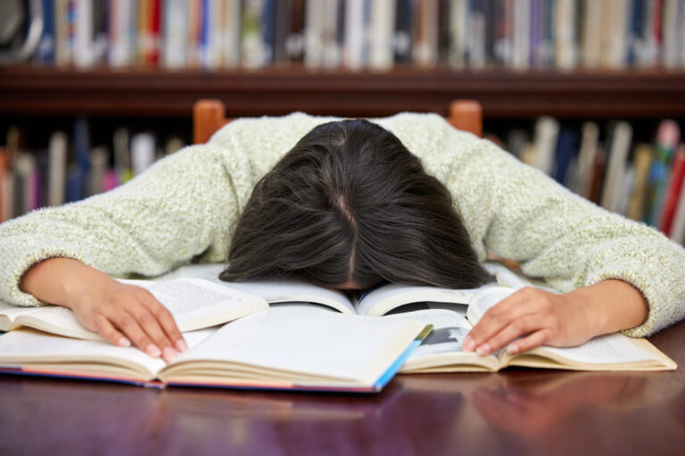 Young woman in a library with her head down on multiple books indicating academic pressure.