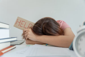 Young woman student holding sign that says “HELP” while surrounded by books and papers indicating the help students need with back to school anxiety.