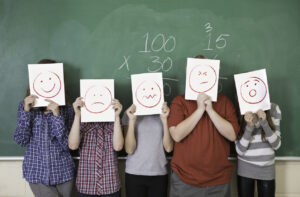 5 students in front of a chalkboard holding papers with various faces smiling, confused, unhappy indicating that parents should look for behavioral changes in their children when they go back to school