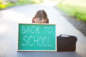 Young white girl with hands covering her face behind a chalk board that says “Back to School” representing the anxiety that children often feel when to go back to school.