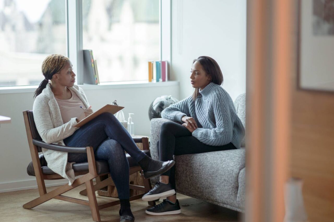 A female Therapist meets with her female client in her office. The client is seated on a sofa with her arms across her body. The Therapist is seated in a chair in front of her as she talks about her client’s mental health.