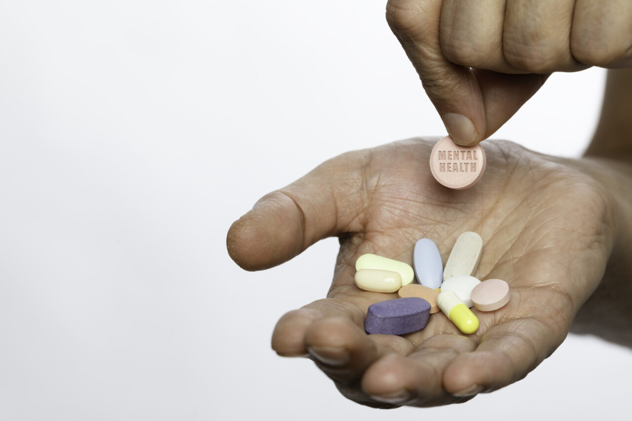 A close-up shot of a hand holding various colorful pills, with one prominently displaying the words "Mental Health." The image emphasizes themes of medication, mental health treatment, and wellness. The white background provides a clean and neutral setting. One hand only is visible in the photo.