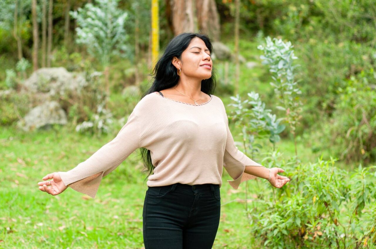 Image of a woman standing at the edge of a forest with her arms outstretched, eyes closed, and a peaceful expression on her face. She seems to be feeling the peace and relief from anxiety that being in nature can bring.
